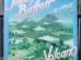 Jimmy Buffett's Volcano album cover featuring illustrated green mountains, a smoking volcano, tropical foliage, and a blue sky with scattered clouds. The album title "Volcano" is displayed in pink and white letters, with "Jimmy Buffett" prominently written at the top in a bold, stylized font. The vinyl cover is positioned in front of a shelf filled with records. Let me know if you’d like any modifications! 🎶🌋
