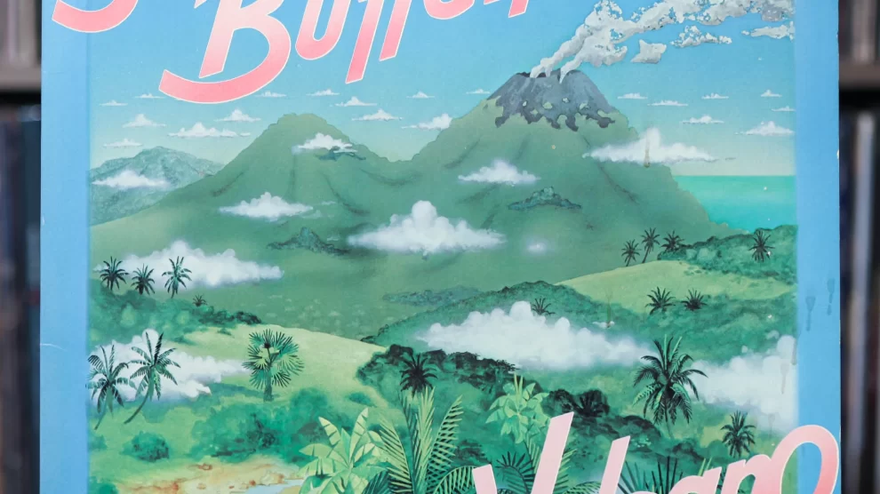 Jimmy Buffett's Volcano album cover featuring illustrated green mountains, a smoking volcano, tropical foliage, and a blue sky with scattered clouds. The album title "Volcano" is displayed in pink and white letters, with "Jimmy Buffett" prominently written at the top in a bold, stylized font. The vinyl cover is positioned in front of a shelf filled with records. Let me know if you’d like any modifications! 🎶🌋