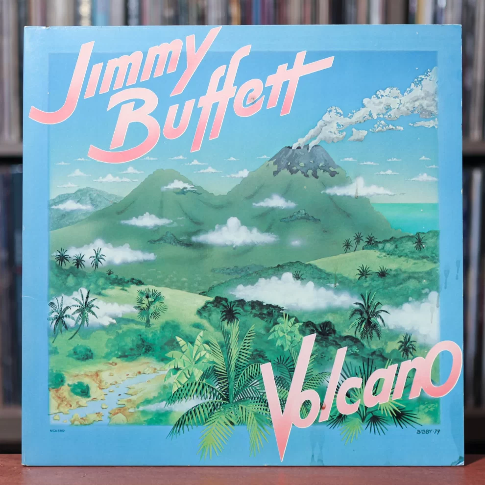 Jimmy Buffett's Volcano album cover featuring illustrated green mountains, a smoking volcano, tropical foliage, and a blue sky with scattered clouds. The album title "Volcano" is displayed in pink and white letters, with "Jimmy Buffett" prominently written at the top in a bold, stylized font. The vinyl cover is positioned in front of a shelf filled with records. Let me know if you’d like any modifications! 🎶🌋